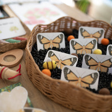 Load image into Gallery viewer, A wicker basket filled with black beans, small orange and yellow beads, and printed butterfly cards arranged on a wooden table in a softly blurred setting.