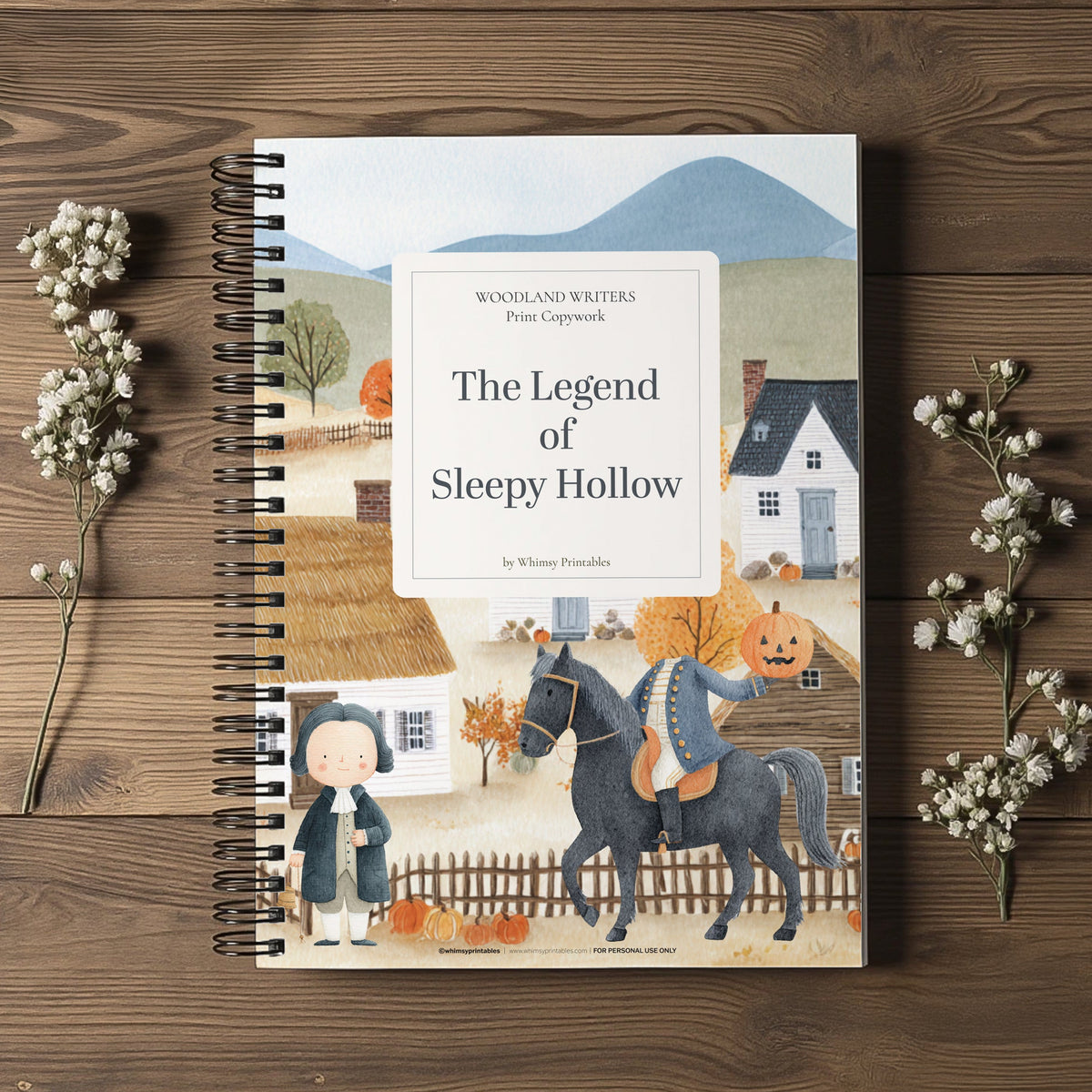 A spiral-bound children's book titled "The Legend of Sleepy Hollow" by Whitney Punshon lies on a rustic wooden surface, styled with dried white flowers on both sides. The cover features a whimsical illustration of a figure on a horse with a pumpkin head and a ghostly figure standing nearby.