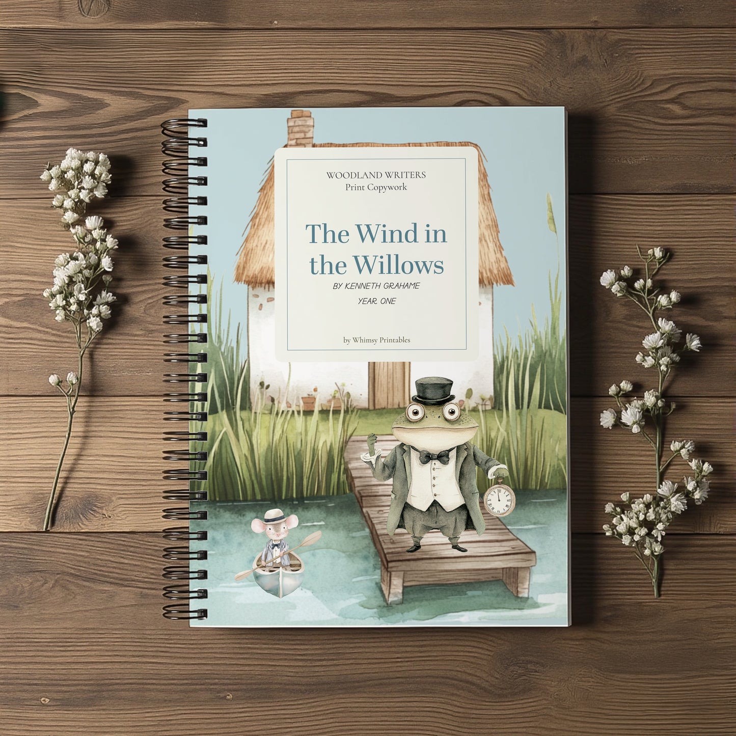 A spiral-bound book titled "The Wind in the Willows" lies flat on a wooden surface, surrounded by small white flower sprigs. The cover shows an illustrated scene with a frog on a dock, reeds, water, and a house.