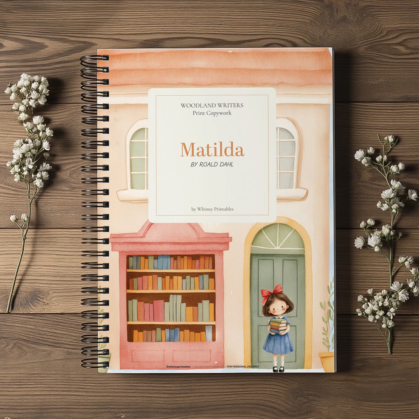 A spiral-bound book titled 'Matilda' by Roald Dahl, featuring a colorful illustration of a girl standing in front of a bookshelf and green door, displayed on a dark wood background with small white flower sprigs.