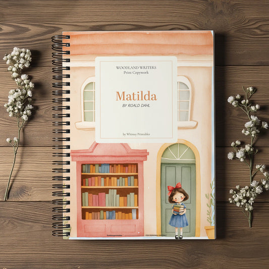 A spiral-bound book titled 'Matilda' by Roald Dahl, featuring a colorful illustration of a girl standing in front of a bookshelf and green door, displayed on a dark wood background with small white flower sprigs.