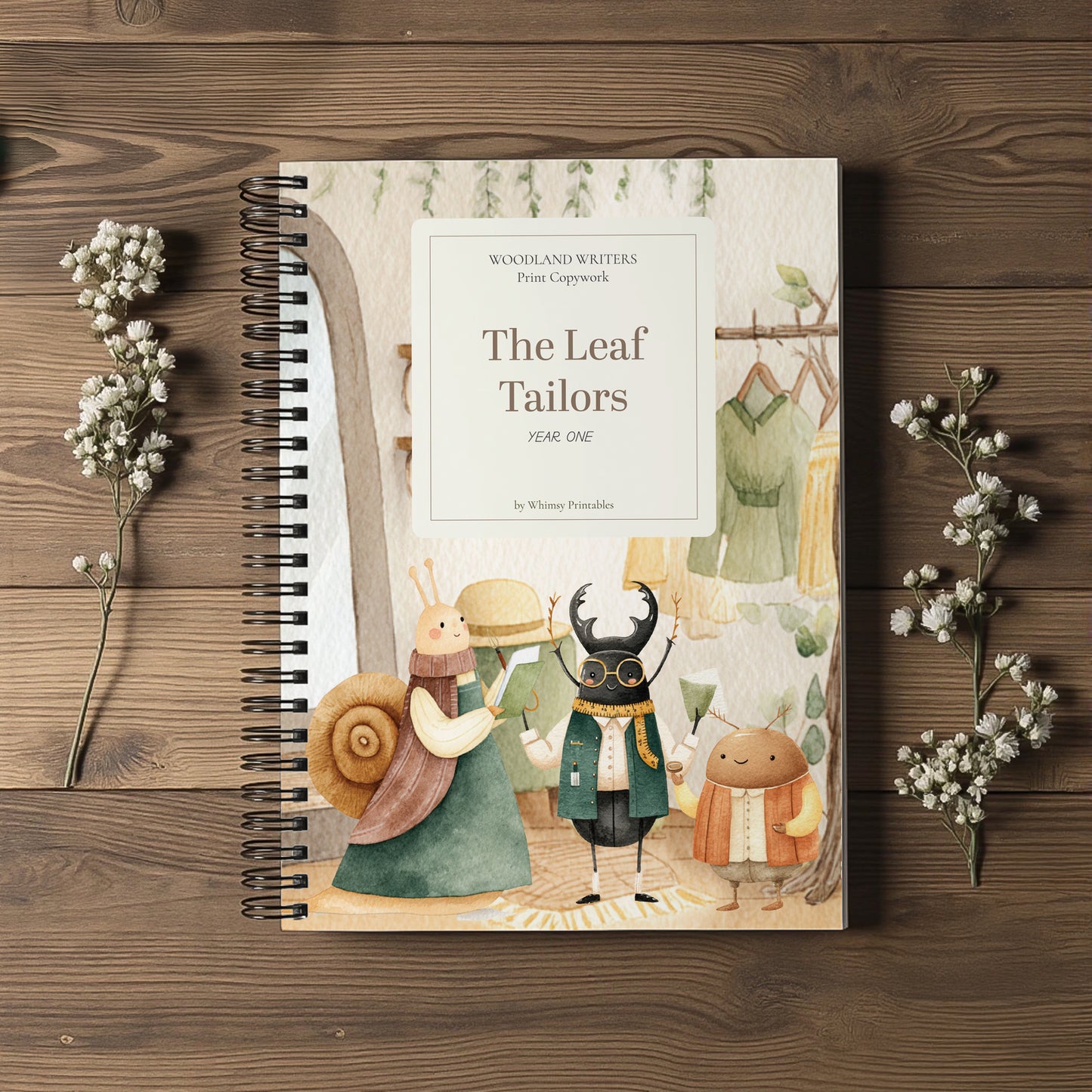 A spiral-bound children's book titled "The Leaf Tailors" by Whitney Penningtons, featuring whimsical illustrated characters on the cover, styled on a dark wood background with small white flowers.