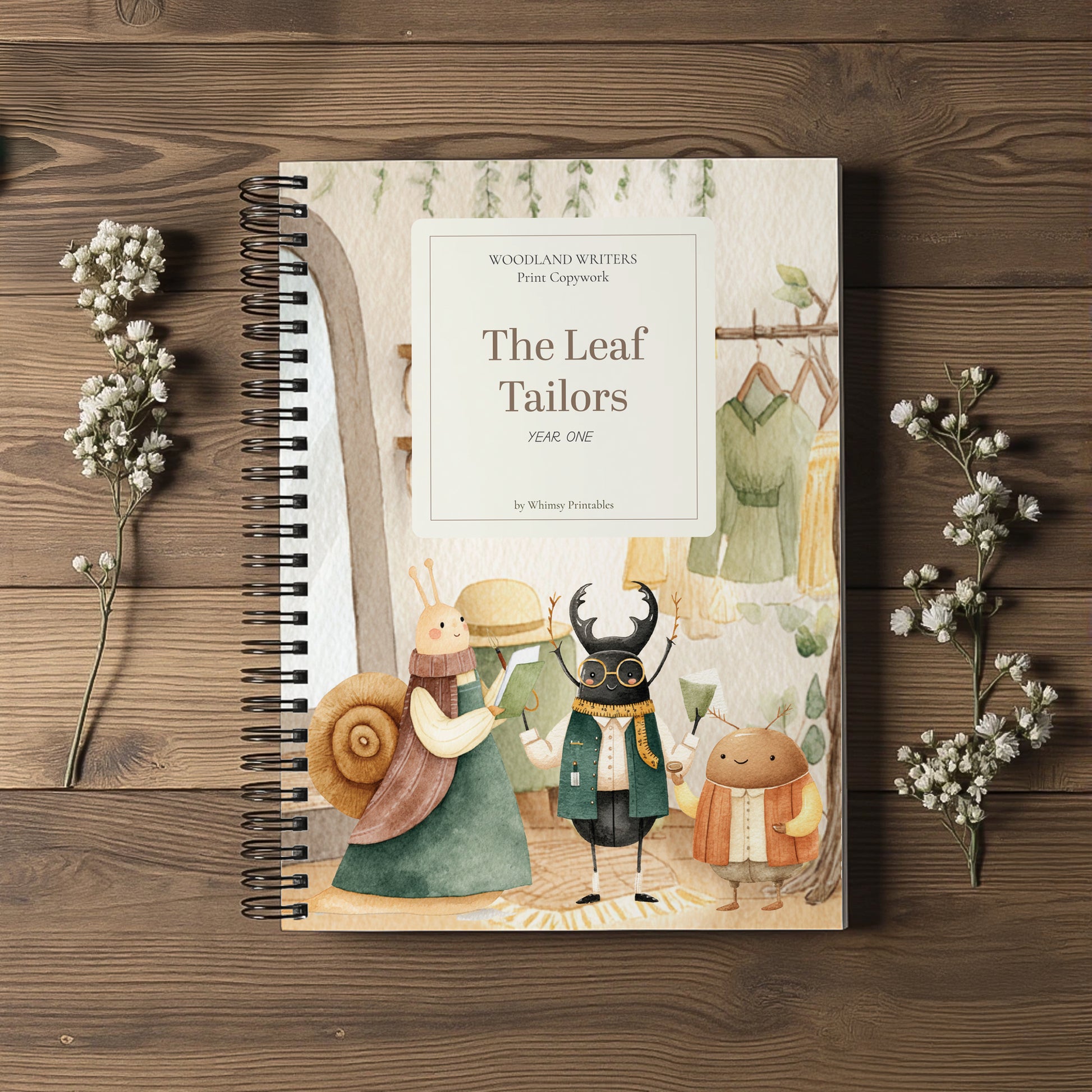 A spiral-bound children's book titled "The Leaf Tailors" by Whitney Penningtons, featuring whimsical illustrated characters on the cover, styled on a dark wood background with small white flowers.