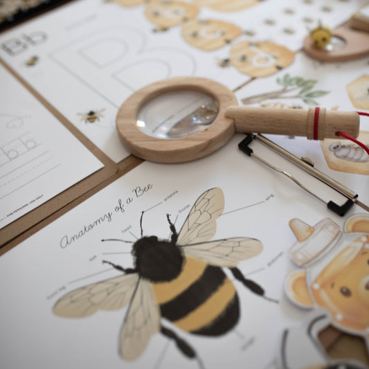 A close-up, angled view of a bee-themed educational activity kit on a white table. The kit includes printed worksheets with bee illustrations, wooden lettering trays, a magnifying glass, tweezers, and stickers.