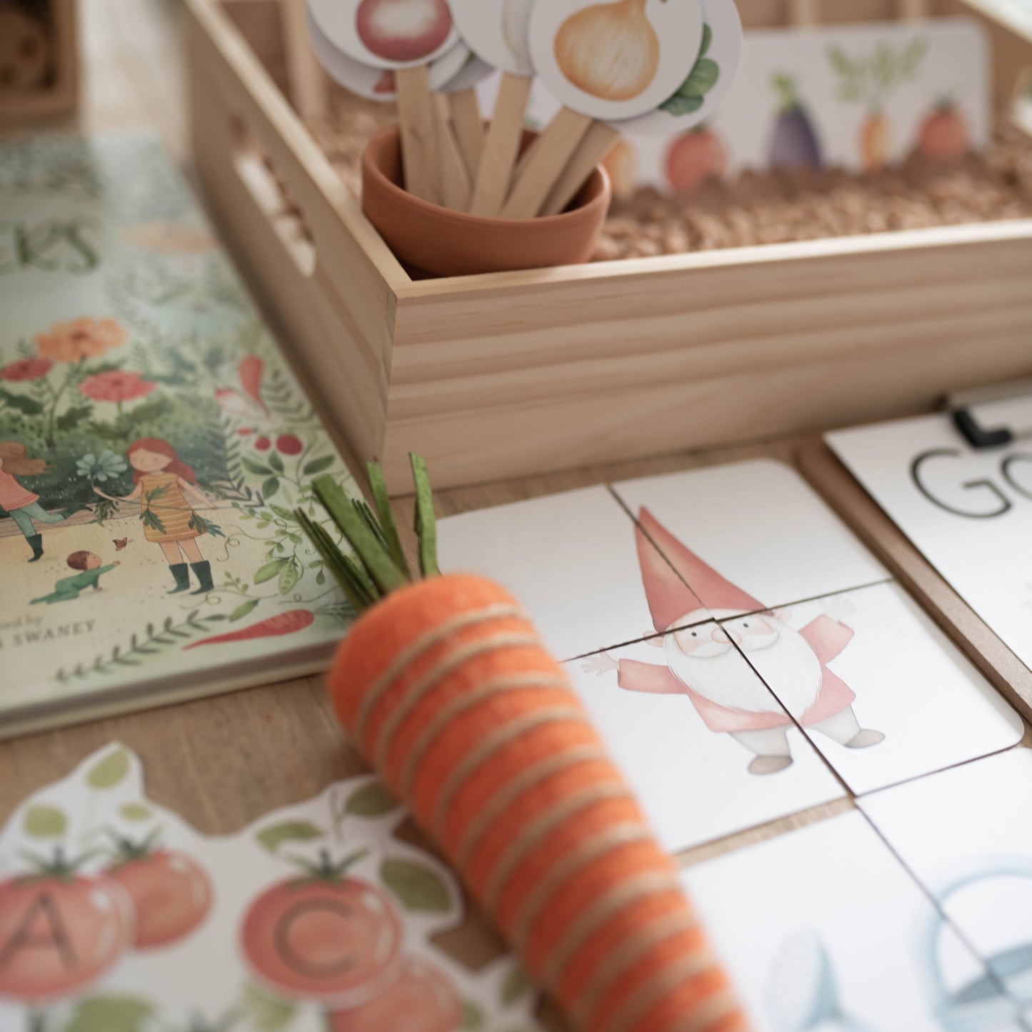 A carrot-shaped orange and beige striped toy or prop lies diagonally across a children's activity surface with illustrated cards, a wooden tray, and small sign pieces in a softly lit indoor setting.