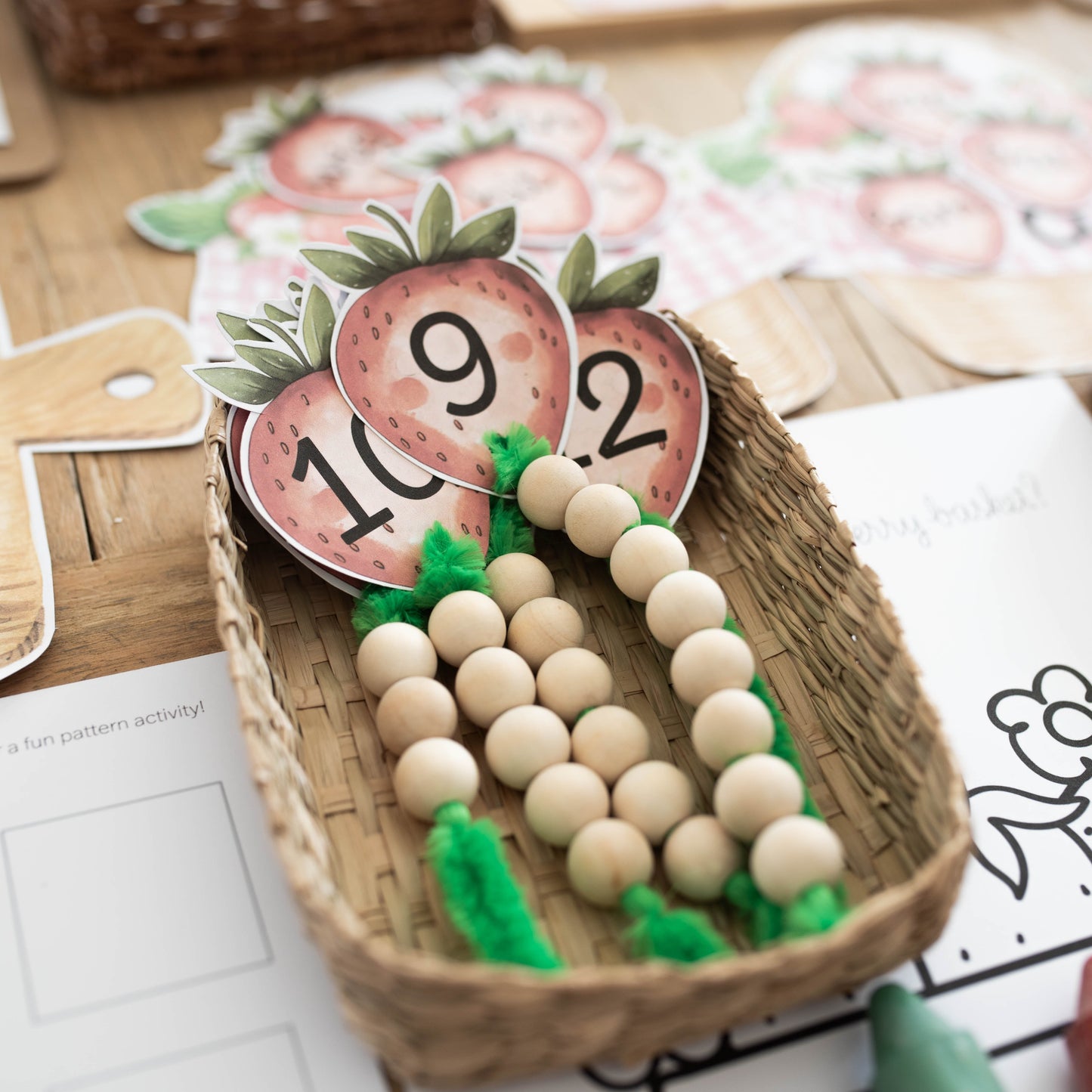 A close-up of a wooden bead counting toy arranged in a small woven basket, with printed number cards and activity sheets on a wooden tabletop.