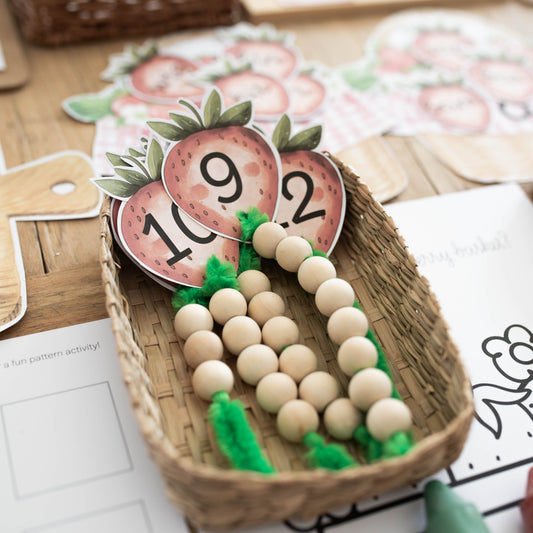 A close-up of a wooden bead counting toy arranged in a small woven basket, with printed number cards and activity sheets on a wooden tabletop.