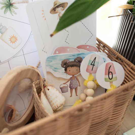 A woven wicker basket filled with children's sensory or educational toys, including illustrated cards, numbered beads, shells, and small accessories, photographed in a bright indoor setting.