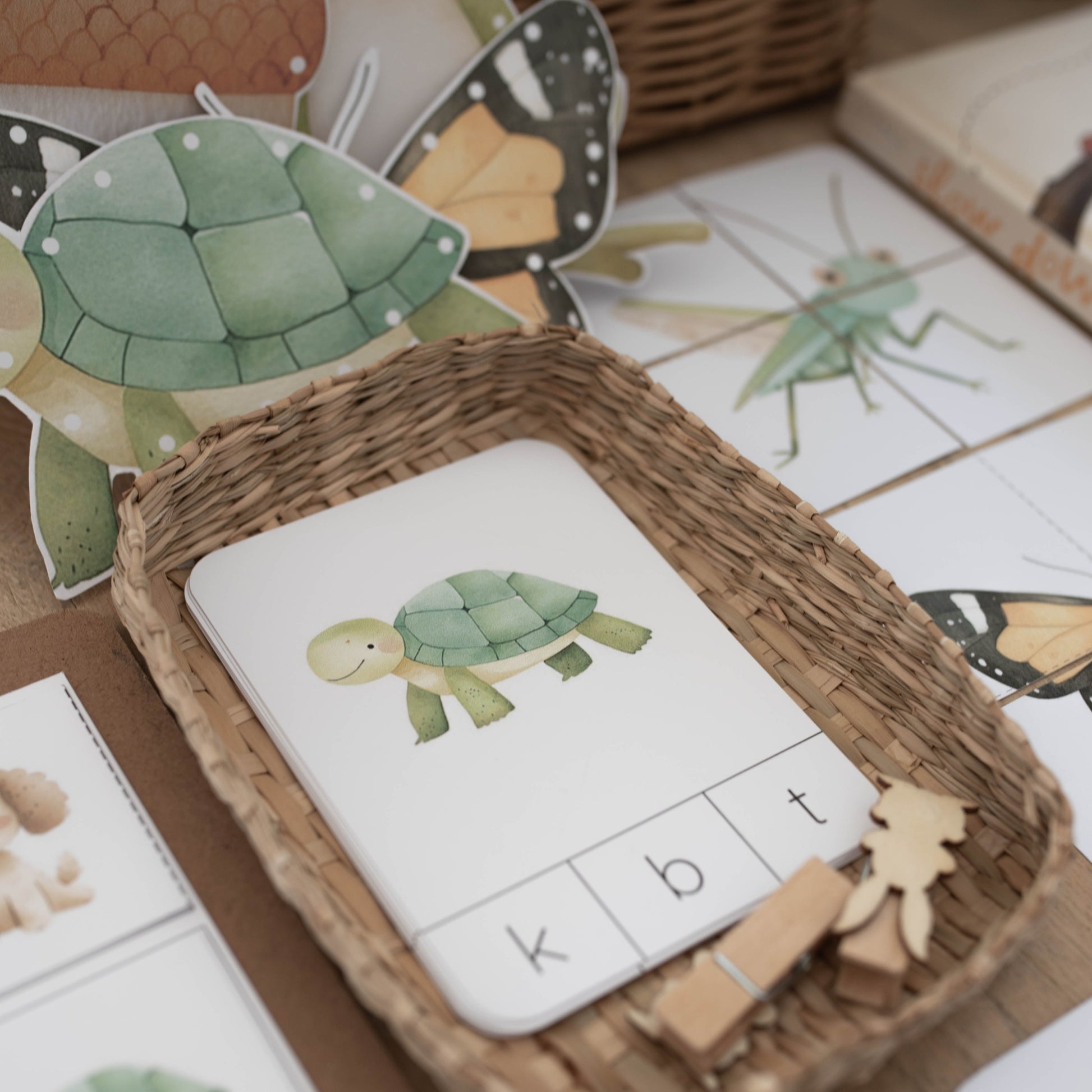 A close-up of a children's educational activity tray made of woven wicker, holding a white card with a green turtle illustration and letters for spelling. Surrounding the tray are matching turtle and insect cards, wooden pegs, and butterfly and turtle cutouts on a tabletop.