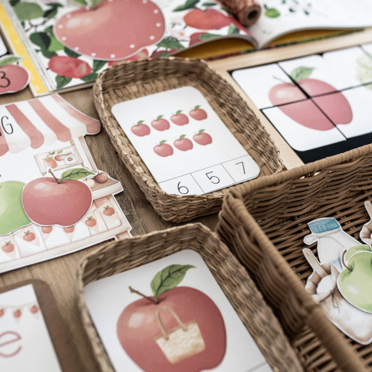 A close-up of children's educational play materials featuring red apples, numbers, and letters, arranged in woven baskets on a wooden surface.