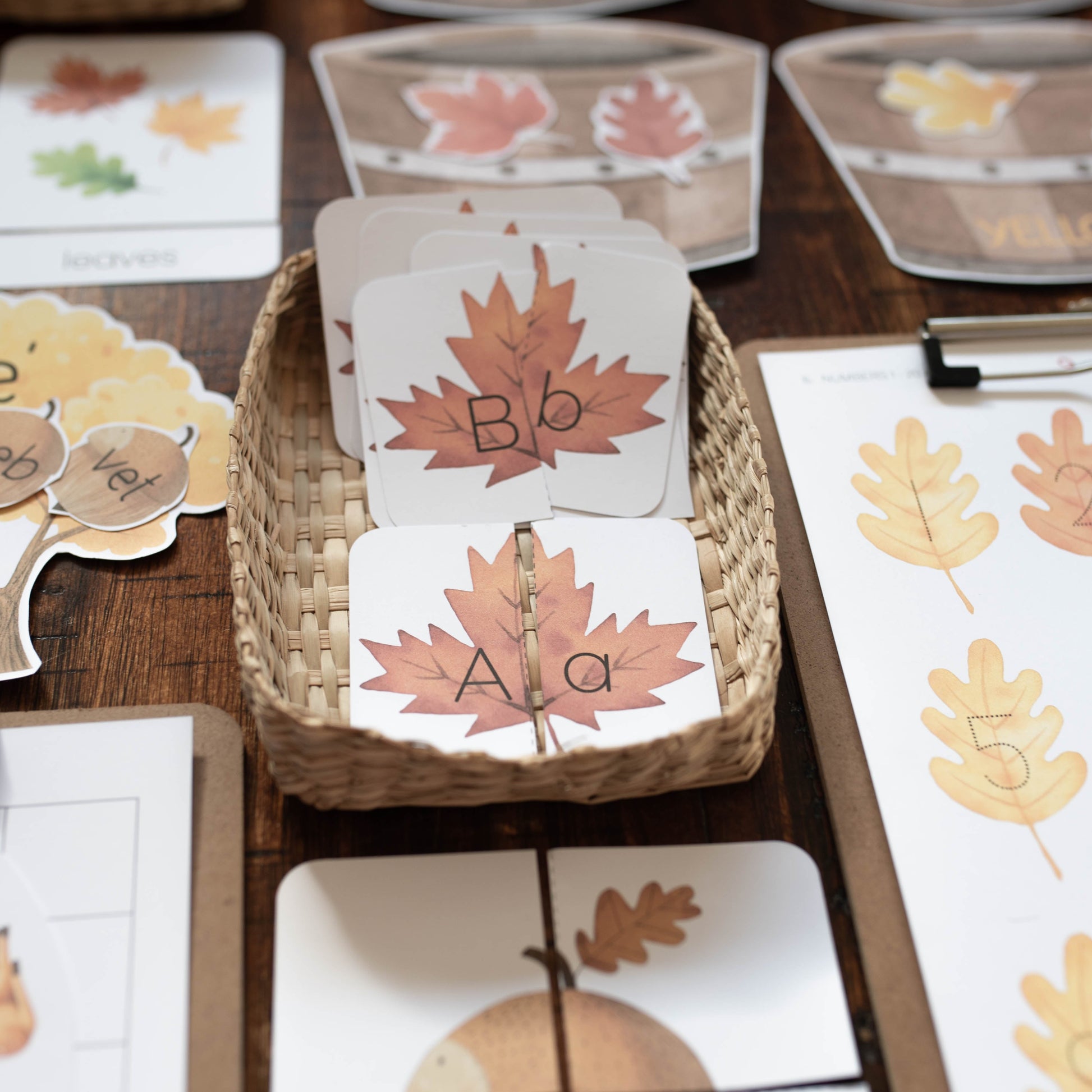 Autumn-themed educational matching cards and visual aids arranged on a wooden table, including leaf cards, acorn cards, and number cards in woven baskets and on a clipboard.