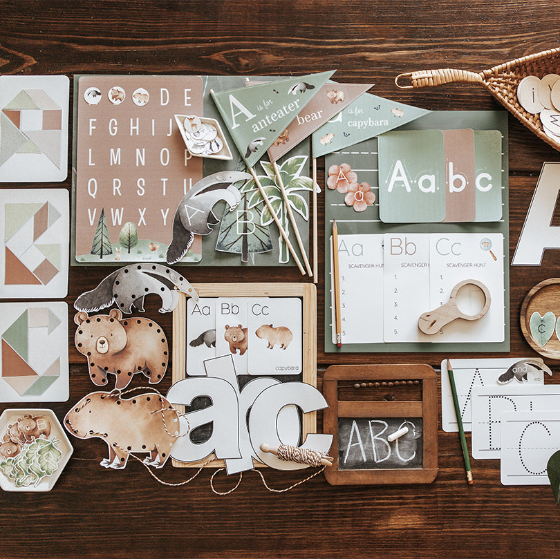 A digital product display featuring a wicker basket with various printable alphabet learning materials, such as flashcards, a chalkboard with letters, and other educational sheets arranged inside and on top of the basket.