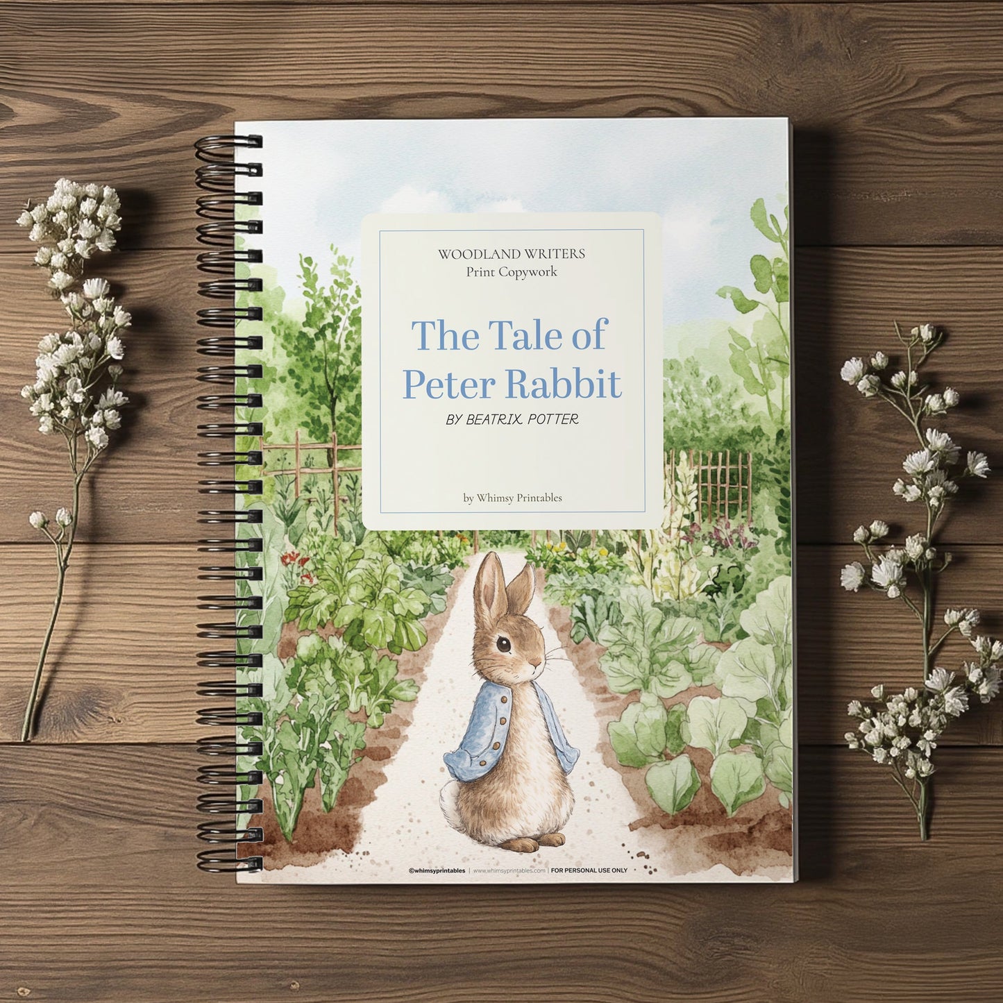 A spiral-bound book titled "The Tale of Peter Rabbit" by Beatrix Potter, photographed from above on a rustic wooden surface with small white flower sprigs placed on both sides. The cover shows a watercolor garden scene with a rabbit wearing a blue bow tie.
