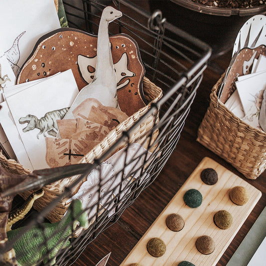 Printed educational activity sheets and materials arranged in a basket, themed around dinosaurs and paleontology, including coloring sheets, matching cards, and lacing cards.